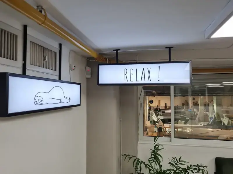 Dos cajas de luz LED colgadas del techo en un pasillo de oficina, una dice RELAX y otra con un dibujo de perezoso